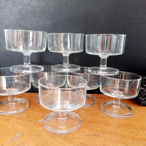 Könnte beinhalten: Ein Set aus sieben Dessertschalen aus klarem Glas, die jeweils eine breite, flache Schale, einen kurzen Stiel und einen runden Fuß aufweisen. Die Schalen sind in einer Gruppe angeordnet, wobei einige vorne stehen und andere im Hintergrund erhöht sind.