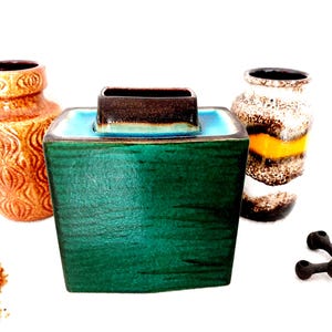 Pode incluir: Uma coleção de vasos de cerâmica vintage em várias formas e cores. A peça central é um vaso quadrado, verde-azulado, com uma parte superior castanha. Outros vasos incluem um vaso laranja texturizado e um vaso castanho e amarelo salpicado. Um objeto decorativo de metal escuro também está presente.