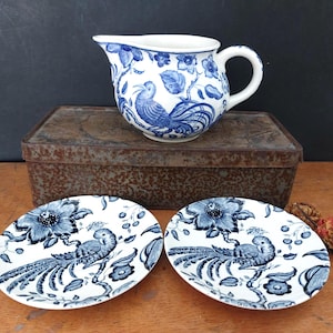 Può includere: Una lattiera in ceramica bianca e due piattini abbinati, tutti decorati con un motivo blu a trasferimento raffigurante un uccello dalla coda lunga appollaiato su un ramo fiorito. Il set è disposto su una scatola di metallo rettangolare.