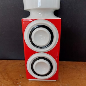 Może przedstawiać: Prostokątny, czerwono-biały ceramiczny wazon o geometrycznym wzorze. Wazon posiada dwa białe okręgi z czarnymi obrysami na czerwonym korpusie. Górna część wazonu jest biała.