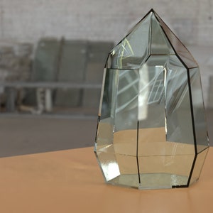May include: A clear, faceted glass terrarium with a geometric design. The terrarium has a pointed top and is displayed on a light brown surface. The background is a blurred industrial setting.