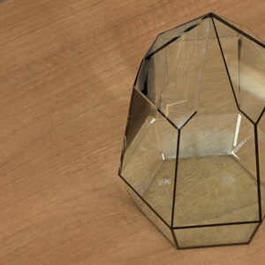 May include: A geometric terrarium with a clear glass design and black trim. The terrarium has multiple facets and is empty. The object is placed on a light brown wooden surface.