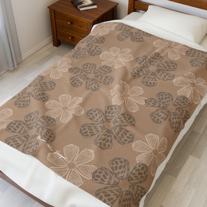 May include: A brown floral patterned blanket with a white border. The blanket features a repeating pattern of stylized flowers in shades of brown and beige. The blanket is on a bed with a white pillow.