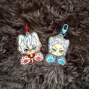 May include: Two acrylic keychains featuring anime-style characters with cat-like features. One keychain has a red and white color scheme, the other blue and white. Both have paw-shaped details and attached key rings.