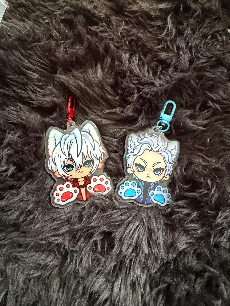 May include: Two acrylic keychains featuring anime-style characters with cat-like features. One keychain has a red and white color scheme, the other blue and white. Both have paw-shaped details and attached key rings.