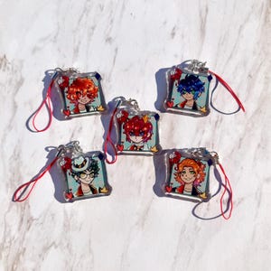 May include: Five square acrylic charms featuring illustrated anime-style characters with vibrant hair colors, each attached to a red phone strap. The designs include red hearts, roses, and small crowns, with silver metal hardware connectors.