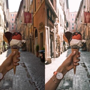 May include: A hand holds a waffle cone ice cream with three scoops: strawberry, vanilla, and chocolate. The ice cream is topped with chocolate sauce and sprinkles. The background is a narrow, cobblestone street in a European city.