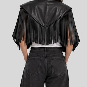 Puede incluir: Una capa corta de cuero negro con un dise&ntilde;o trasero en punta y largos flecos colgantes a lo largo de los bordes. La prenda se lleva sobre una camisa blanca lisa y se combina con pantalones vaqueros de mezclilla gris oscuro, vista trasera.