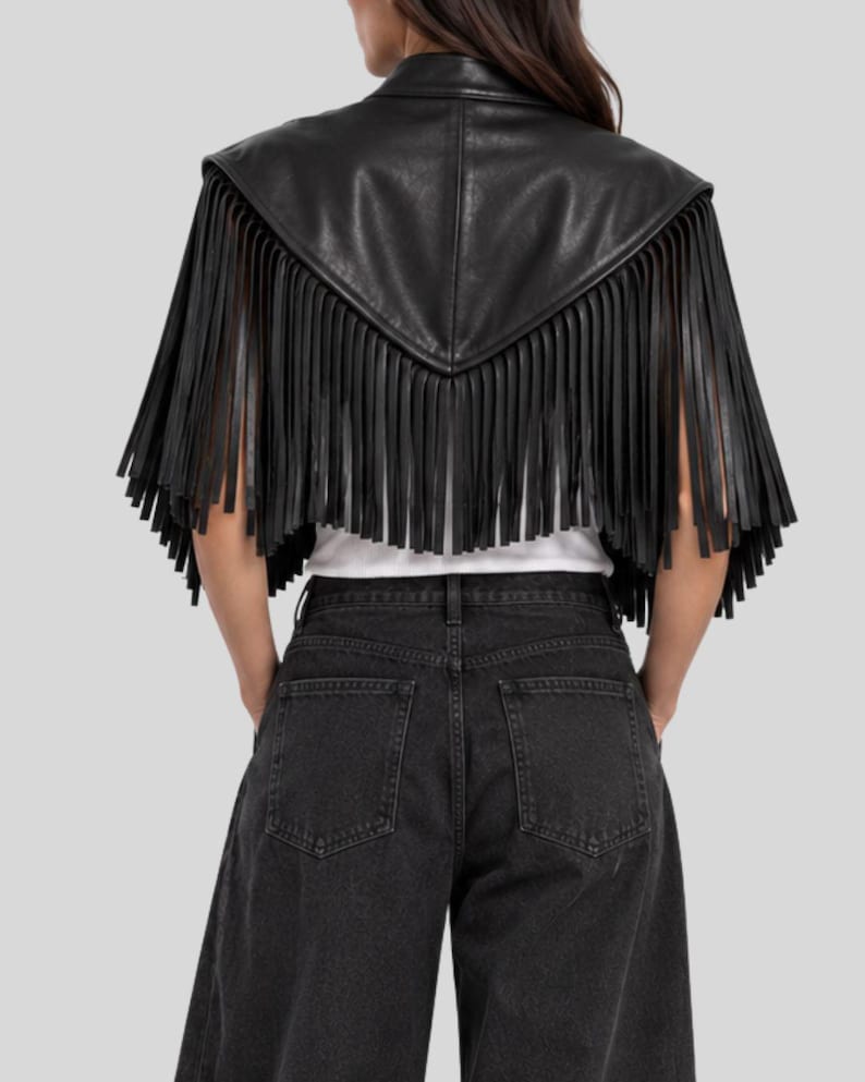 Puede incluir: Una capa corta de cuero negro con un dise&ntilde;o trasero en punta y largos flecos colgantes a lo largo de los bordes. La prenda se lleva sobre una camisa blanca lisa y se combina con pantalones vaqueros de mezclilla gris oscuro, vista trasera.