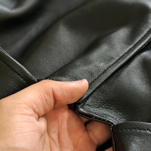 May include: Close-up of black leather material, showing the texture and stitching. The leather appears smooth and has a slight sheen. The image highlights the craftsmanship and detail of the material.