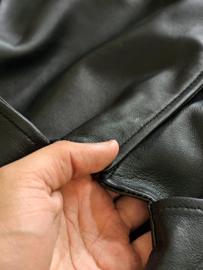 May include: Close-up of black leather material, showing the texture and stitching. The leather appears smooth and has a slight sheen. The image highlights the craftsmanship and detail of the material.