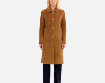 May include: A tan suede coat featuring a collared neckline, long sleeves, and a front button closure with four visible buttons. The coat has a straight silhouette that extends to the mid-thigh, paired with dark navy blue trousers and black shoes.