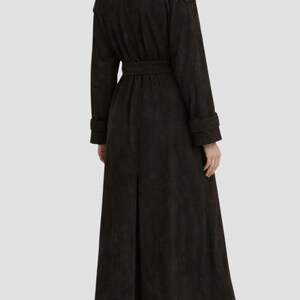 May include: A long, dark brown trench coat featuring a collared neckline, long sleeves with cuff straps, and a matching tie belt at the waist. The coat has a structured shoulder design and a single back vent, extending down to the lower calf area.