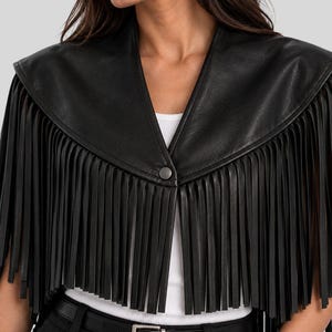 Puede incluir: Una capa corta de cuero negro con un amplio escote en V, un cierre de bot&oacute;n a presi&oacute;n central y largos flecos rectos que se extienden desde la l&iacute;nea de los hombros hasta el dobladillo, sobre una camiseta blanca sin mangas y pantalones negros.