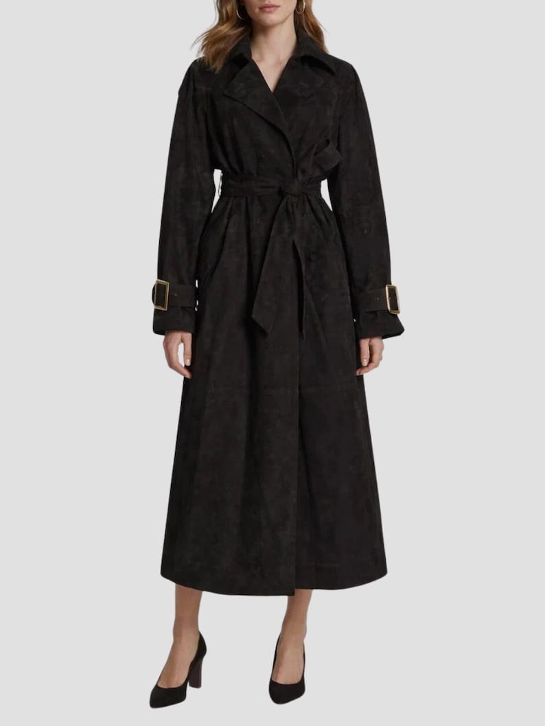 May include: A long, black trench coat featuring a wide notched collar, long sleeves with buckled straps at the cuffs, and a matching tie belt at the waist. The coat has a textured finish and falls to a midi length, providing a structured silhouette.