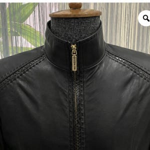 Könnte beinhalten: Eine schwarze Jacke mit Stehkragen und einem mittigen, goldfarbenen Reißverschluss mit rechteckigem Zipper. Das Kleidungsstück weist strukturierte Details an den Schultern und entlang der Vorderseite auf, die sich vom glatten Material abheben.