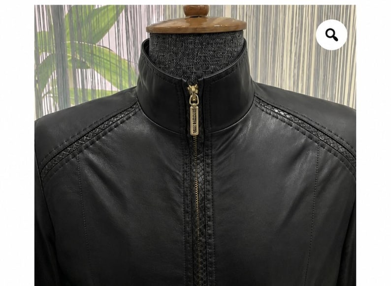 May include: A black jacket with a high collar and a central gold-toned zip featuring a rectangular pull tab. The garment includes textured detailing along the shoulders and down the front placket, contrasting with the smooth black material.
