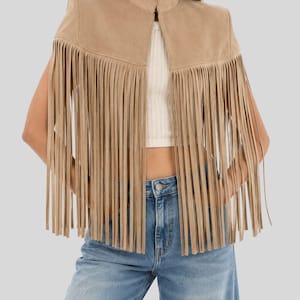 Puede incluir: Una chaqueta de ante color canela con cuello alto y largos flecos fluidos que se extienden desde el pecho hasta la cintura. La chaqueta se lleva sobre un top corto de canalé blanco y vaqueros azules, destacando un diseño de hombros estructurados.