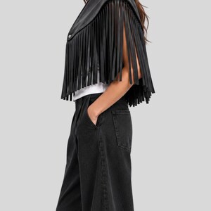 Puede incluir: Una capa corta de cuero negro con cuello ancho, cierre de un solo bot&oacute;n en la parte delantera y flecos largos y finos que se extienden desde los hombros hasta el dobladillo, sobre una camiseta blanca lisa y pantalones vaqueros negros.