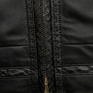 May include: A black jacket featuring a central metal zip closure. The garment is constructed with smooth black panels accented by horizontal strips of textured, snakeskin-patterned leather. Decorative stitching lines border the textured sections.