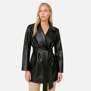 Puede incluir: Un abrigo tipo trench negro de cuero sintético con cinturón en la cintura. El abrigo tiene un cuello con solapas clásico y mangas largas. La modelo lleva el abrigo con pantalones color beige.