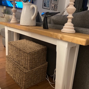 May include: A wooden console table with a light wood top and white legs, with two woven storage baskets underneath. A white jug, lamps, and decorative items are on the table. The table is against a grey sofa.