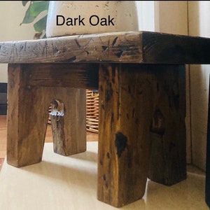 May include: A dark oak wood stool with a rustic, distressed finish. The stool has a simple design with four legs and a rectangular top. The text "Dark Oak" is visible on the top of the stool.