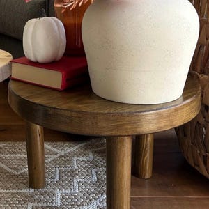 May include: A round wooden side table with three legs. The table is decorated with a white ceramic vase with a pink rose and a white pumpkin.