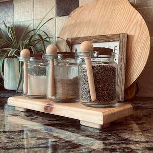 May include: Three glass jars with wooden spoons, filled with white, brown, and black spices, sitting on a wooden tray. The jars are on a worktop with a wooden chopping board and a framed sign in the background.