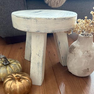 May include: A small, round, white-washed wooden stool with three legs. The stool has a distressed finish, and is next to a white vase with dried flowers and two gold pumpkins. The stool is approximately 25 cm in diameter.