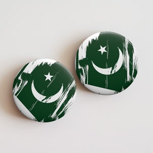 May include: Two round, glossy badges featuring the flag of Pakistan. The design includes a white crescent moon and star on a dark green background, with white brushstroke accents. The badges are displayed on a white surface.