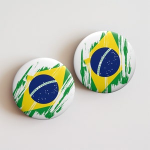 May include: Two round white buttons featuring the Brazilian flag design. The flag's elements include a blue circle with stars, a yellow diamond, and green brushstrokes. The text "ORDEM E PROGRESSO" is visible on the blue circle.