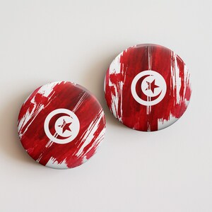 May include: Two round buttons featuring the Tunisian flag. The design includes a white crescent moon and star on a red background with white brushstrokes. The buttons are displayed on a white surface.