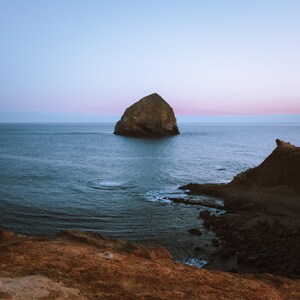 May include: A scenic coastal landscape featuring a large rock formation in the ocean. The water is a deep blue, and the sky transitions from pink to blue. Rugged cliffs frame the scene, creating a serene and natural environment.