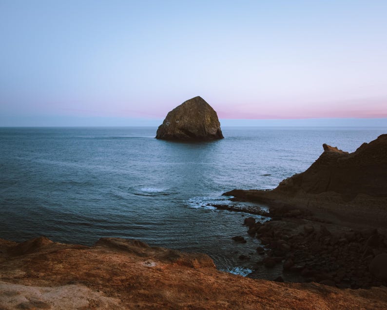 May include: A scenic coastal landscape featuring a large rock formation in the ocean. The water is a deep blue, and the sky transitions from pink to blue. Rugged cliffs frame the scene, creating a serene and natural environment.
