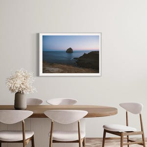 May include: A framed landscape photograph of a coastal scene. The image features a large rock formation in the ocean under a dusky blue sky. The photograph is displayed on a white wall above a dining table with chairs and a vase of dried flowers.