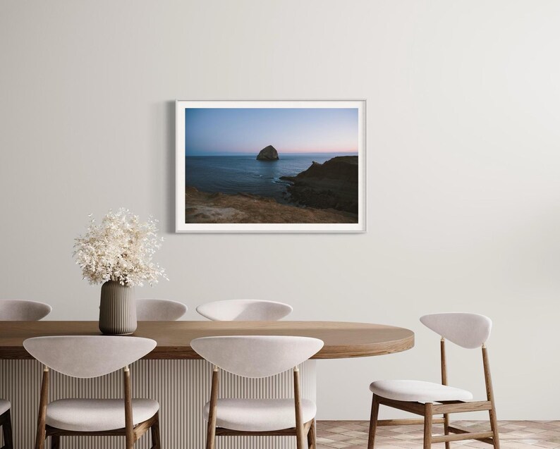 May include: A framed landscape photograph of a coastal scene. The image features a large rock formation in the ocean under a dusky blue sky. The photograph is displayed on a white wall above a dining table with chairs and a vase of dried flowers.