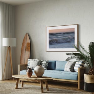 May include: A living room interior with a blue sofa, a wooden coffee table, a surfboard leaning against the wall, a potted palm tree, and a framed photograph of ocean waves.