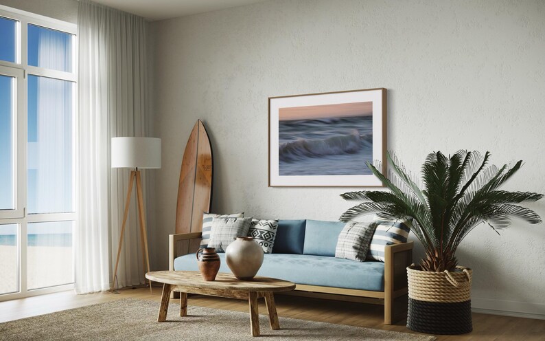 May include: A living room interior with a blue sofa, a wooden coffee table, a surfboard leaning against the wall, a potted palm tree, and a framed photograph of ocean waves.