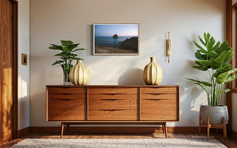 May include: A mid-century modern style room with a wooden credenza, two decorative vases, and a framed seascape painting. A large potted plant and a smaller plant in a glass vase add greenery. A gold wall sconce is mounted on the wall.