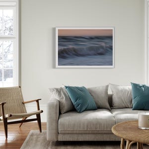 May include: A framed photograph of a wave crashing on a beach at sunset. The photo is in a white frame and is hanging on a white wall. The photo is in a living room setting with a couch, coffee table, and two chairs.