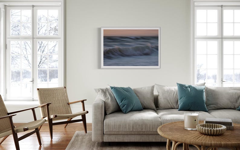 May include: A framed photograph of a wave crashing on a beach at sunset. The photo is in a white frame and is hanging on a white wall. The photo is in a living room setting with a couch, coffee table, and two chairs.