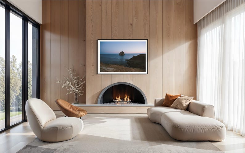 May include: A modern living room with a fireplace, a large framed landscape art piece, and a curved sofa. The room features light-colored walls, a neutral rug, and large windows with sheer curtains. Two chairs are in the foreground.