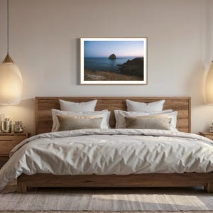 May include: A bedroom scene featuring a large wooden bed with a light-colored duvet and pillows. A framed landscape art piece hangs above the bed. Two pendant lights with a teardrop shape hang on either side of the bed. A potted plant sits on the right.