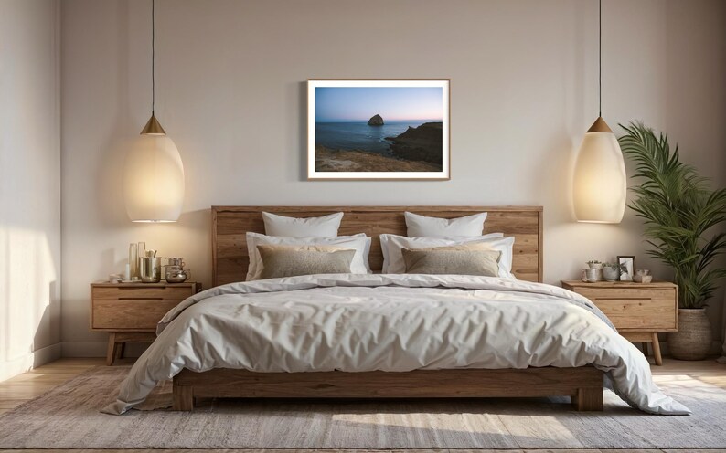 May include: A bedroom scene featuring a large wooden bed with a light-colored duvet and pillows. A framed landscape art piece hangs above the bed. Two pendant lights with a teardrop shape hang on either side of the bed. A potted plant sits on the right.