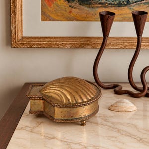 May include: A brass trinket box shaped like a scallop shell with a textured, ribbed lid and a rope-style border. The box features a weathered, metallic gold finish and rests on small, rounded feet, accompanied by a small, natural seashell.