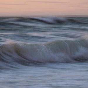 May include: A close-up of a wave crashing on the shore, creating a soft, blurred effect. The water is a light blue and white, with a hint of pink in the sky.