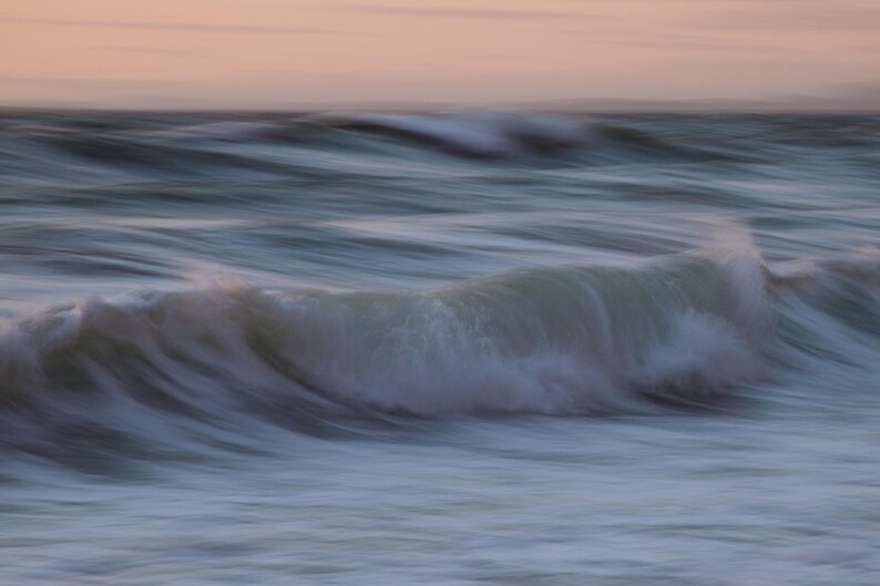 May include: A close-up of a wave crashing on the shore, creating a soft, blurred effect. The water is a light blue and white, with a hint of pink in the sky.