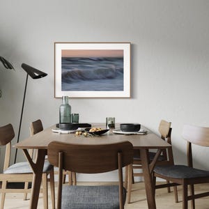 May include: A framed photograph of a blurry ocean wave with a light blue, white, and pink color scheme hangs on a white wall above a wooden dining table with four chairs.