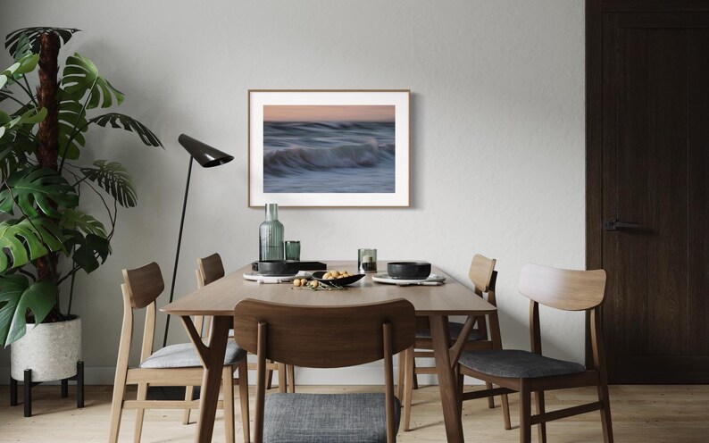 May include: A framed photograph of a blurry ocean wave with a light blue, white, and pink color scheme hangs on a white wall above a wooden dining table with four chairs.